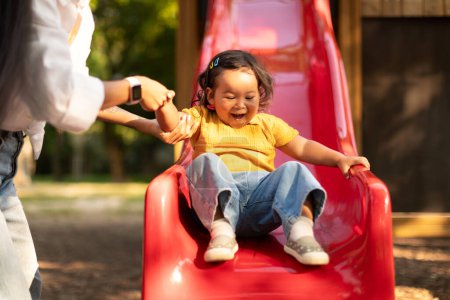 Photo for Asian Mother Catching Her Happy Baby Daughter As Child Riding Slides At Public Playground In City Park Outdoor. Selective Focus On Infant, Cropped. Toddler Girl Having Fun With Mom Playing Outside - Royalty Free Image