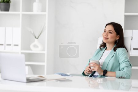 Foto de Calma soñando sonriente dama caucásica milenaria en traje descansando en el lugar de trabajo con una taza de té y computadora, mira el espacio libre en el interior de la oficina. Pausa para café, negocio con dispositivo, trabajo, relajarse - Imagen libre de derechos