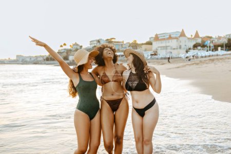 Foto de Tres amigas felices y diversas en traje de baño y sombreros de paja abrazando y disfrutando del tiempo juntas mientras caminan en la playa en la orilla del océano - Imagen libre de derechos
