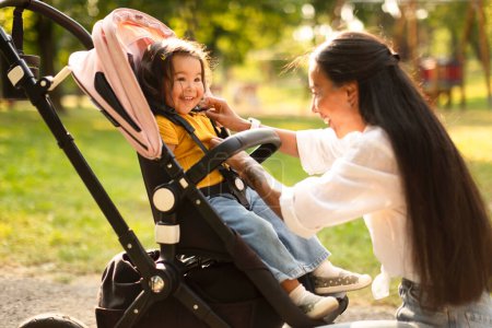Foto de Pasea con Baby. Madre asiática feliz que hace a su hija pequeña cómoda mientras está sentada en el cochecito, mujer ajustando correas de seguridad para comodidad caminando en el parque al aire libre. Enfoque selectivo - Imagen libre de derechos
