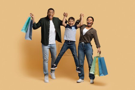 Photo for Funny adorable happy young black family have fun while shopping together isolated on beige background. Loving father and mother holding purchases and lifting up their cute preteen son - Royalty Free Image