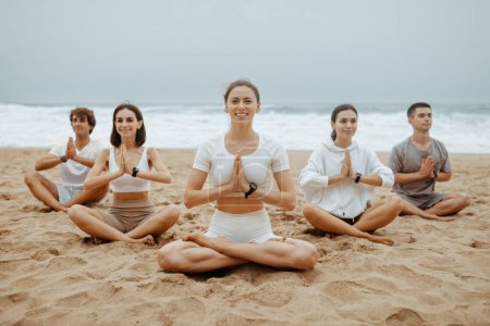 Foto de Jóvenes practican meditación en la playa. Grupo de hombres y mujeres meditando en la orilla del mar, disfrutando de bienestar y estilo de vida saludable, sonriendo a la cámara - Imagen libre de derechos