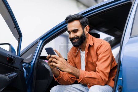Foto de Aplicación de navegación del coche. Hombre de Oriente Medio feliz usando teléfono sentado en auto con la puerta abierta, la búsqueda de la mejor manera a través de Mobile Gps Navigator App, Posando con el gadget en el automóvil de lujo - Imagen libre de derechos