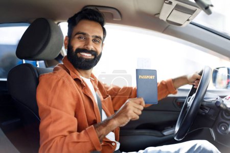 Foto de Viaje en coche. Hombre conductor indio feliz mostrando su pasaporte sentado dentro del automóvil, viajando al extranjero por automóvil moderno, sonriendo a la cámara. Feliz turista disfrutando de paseo en vehículo alquilado en nuevo país - Imagen libre de derechos