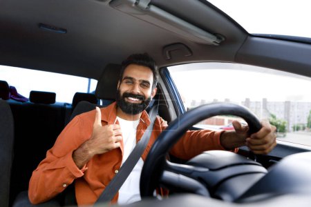 Foto de Concepto de venta al por menor. Hombre árabe feliz en traje casual conduciendo coche de lujo, mostrando el pulgar hacia arriba sentado dentro del vehículo y sonriendo a la cámara disfrutando de paseo a través de un automóvil cómodo a estrenar - Imagen libre de derechos