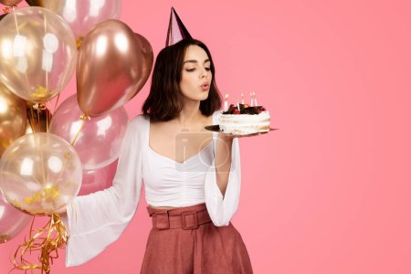 Photo for Positive millennial caucasian brunette lady in hat with many balloons blows out candles on cake, make wish, isolated on pink background, studio. Celebration birthday, holiday party, ad and offer - Royalty Free Image