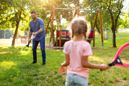 Photo for Back view of little girl holding badminton racket while playing with her father in the park on sunny summer day, family spending free time together outdoors - Royalty Free Image