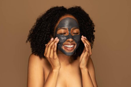Foto de Concepto de cuidado de la piel. Emocionado feliz semidesnuda jovencita negra disfrutando de la nueva máscara orgánica, tocando la máscara limpiadora de carbón en su cara y sonriendo, aislado sobre fondo marrón - Imagen libre de derechos