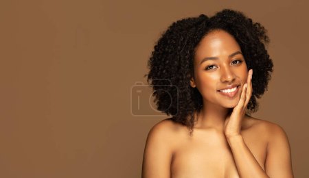 Photo for Face care routine, beauty procedure, aesthetic medicine, cosmetology. Cheerful half-naked african american young woman with bushy hair touching her face and smiling, isolated on brown background - Royalty Free Image