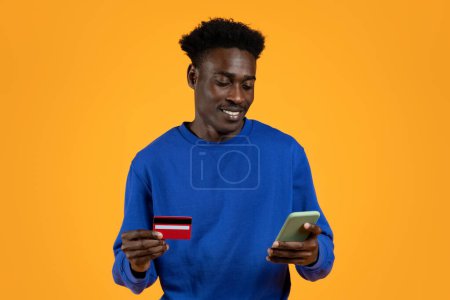 Téléchargez les photos : Heureux jeune homme noir attrayant portant pull bleu avec carte bancaire en plastique rouge et téléphone portable dans ses mains en regardant l'écran du gadget, fond jaune. Shopping, banque en ligne - en image libre de droit