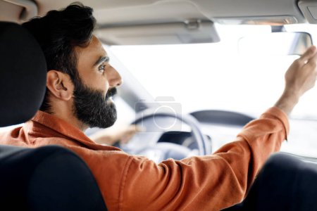 Foto de Automóvil nuevo. Vista trasera del joven indio conduciendo su coche y ajustando el espejo mientras monta en la ciudad. Tiro trasero del dueño contento del automóvil que prueba y que compra el vehículo moderno - Imagen libre de derechos