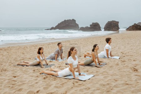 Téléchargez les photos : Tranquillité des marées. Groupe de jeunes hommes et femmes pratiquant le yoga et faisant poser le cobra sur des nattes, faisant de l'exercice sur la plage au bord de l'océan le matin, copiant l'espace - en image libre de droit
