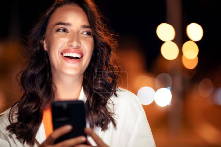 Photo for Womans happy night stroll with her smartphone, lady texting or surfing internet while walking on the street, looking aside and smiling, copy space - Royalty Free Image