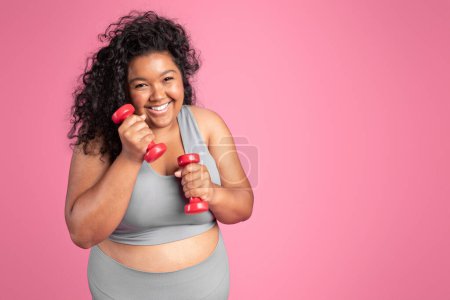 Photo for Excited plus size black woman in sportswear holding dumbbells and laughing, standing on pink studio background, copy space. Workout for body care, weight loss - Royalty Free Image