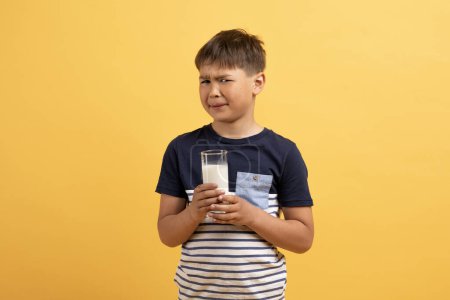Photo for Lactose intolerance for children concept. Unhappy school aged kid boy in casual outfit holding glass of milk in his hands and grimacing, isolated on yellow studio background, copy space - Royalty Free Image