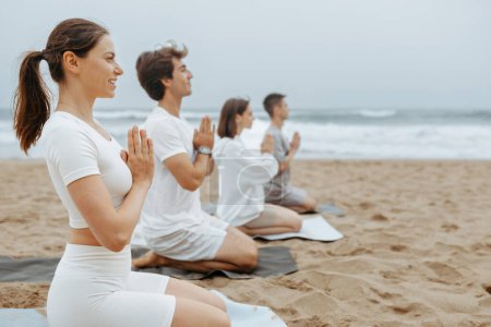Photo for Youths practising yoga and meditation on the beach, sitting on mats with clasped hands, enjoying training outdoors on the beach, side view, free space - Royalty Free Image