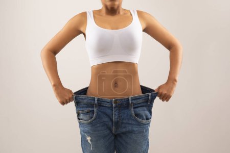 Photo for Cropped of slim well-fit black woman wearing huge pants, showing her big old jeans, demonstrating results of diet, workout, weight loss program, grey background. Slimming concept - Royalty Free Image