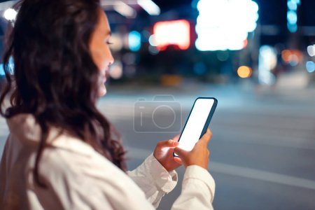 Photo for Young woman using smartphone with blank screen, standing at night city street near road, using new mobile app for navigating in megalopolis, mockup, free space - Royalty Free Image