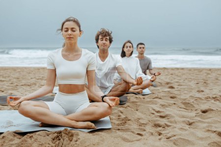 Photo for Oceanfront om. Calm women and men sitting in row in lotus pose holding fingers in gyan mudra hand gesture to maintaining focus, meditating on the beach, copy space - Royalty Free Image
