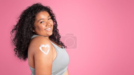 Foto de Rutina diaria de cuidado de la piel. Retrato de mujer negra feliz más tamaño con loción corporal en forma de corazón en el hombro, fondo de estudio rosa, pancarta, panorama, espacio libre - Imagen libre de derechos