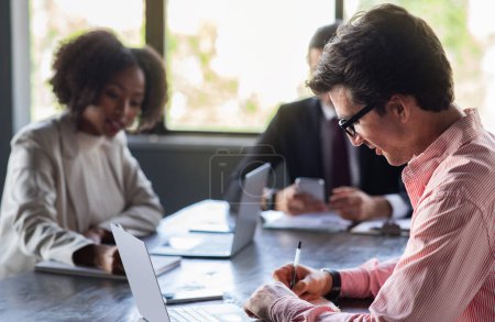 Photo for International management team professionals young black woman and caucasian men have briefing at meeting room at office. Employees sitting at desk at conference hall, using gadgets, taking notes - Royalty Free Image