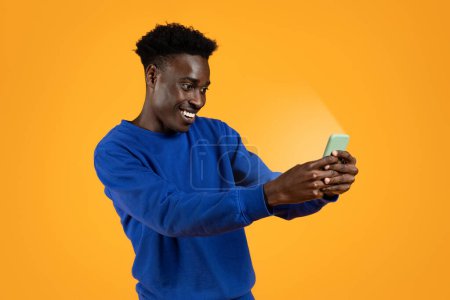 Téléchargez les photos : Offre en ligne incroyable, deal. Excité joyeux beau jeune homme noir regardant l'écran brillant smartphone, fond studio jaune. Heureux homme afro-américain utilisant un téléphone portable, contenu incroyable - en image libre de droit