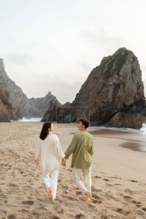 Téléchargez les photos : Plan vertical de jeune couple amoureux marchant sur la plage au bord de l'océan, se tenant la main et appréciant la date et l'heure ensemble, espace libre, pleine longueur - en image libre de droit