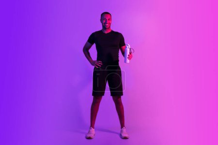 Photo for Healthy sport motivation. Smiling sporty black guy in fitwear holds bottle of water, advertising gym, fitness offer on purple neon background. Full length of positive african sportsman, copy space - Royalty Free Image