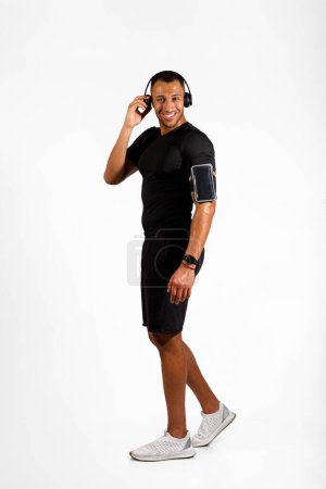 Photo for Workout Playlist. Vertical Shot Of Black Sporty Young Man Wearing Earphones And Looking At Camera, Posing In Fitwear On White Studio Background. Music For Fitness Training. Full Length - Royalty Free Image