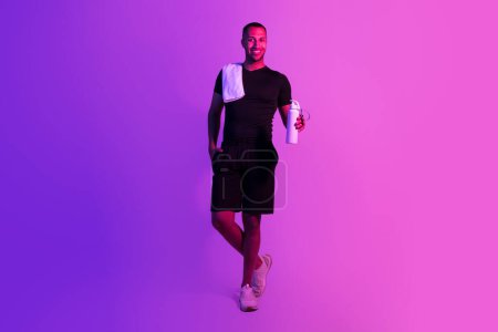 Téléchargez les photos : Plan complet de l'homme de fitness afro-américain en vêtements de sport noirs dans la lumière pourpre néon, tient bouteille d'eau et serviette prête pour l'entraînement dans la salle de gym, debout sur le fond du studio, souriant à la caméra - en image libre de droit