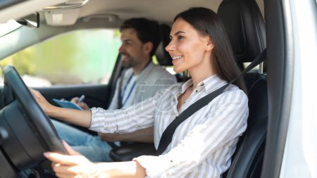 Photo for Cheerful confident pretty young woman sitting inside car with man instructor by her side, lady student holding hands on steering wheel, passing exams successfully at driving school, getting licence - Royalty Free Image