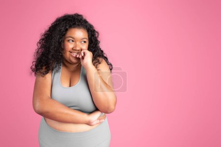 Photo for Young african american obese woman in sports outfit posing and looking aside at free space, standing on pink studio background. Active lifestyle and weight loss concept - Royalty Free Image