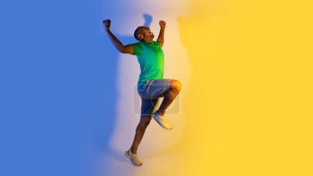 Photo for Victory and achievement. Emotional african american sporty guy running, winner shouting celebrating his sport triumph, spreading hands and shaking fists over blue and yellow background. Panorama - Royalty Free Image