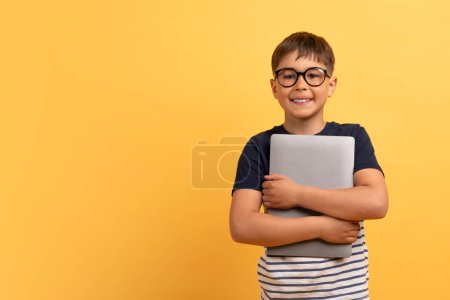 Photo for Cheerful handsome cute guy school aged kid boy in casual t-shirt and eyeglasses embracing computer laptop and smiling, isolated on yellow background, copy space. Gadget addiction in children - Royalty Free Image