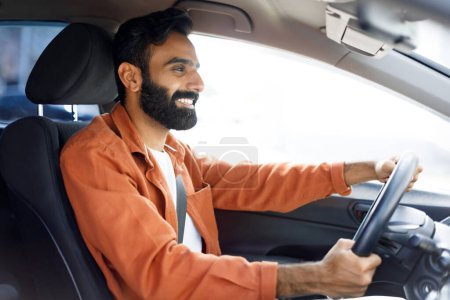 Foto de Auto nuevo. Hombre indio sonriente conduciendo disfrutando de la comodidad de un nuevo vehículo moderno, sentado en el asiento del conductor en el automóvil. Vista lateral tiro de conductor chico posando sosteniendo rueda dentro del coche - Imagen libre de derechos