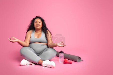 Téléchargez les photos : Corps noir paisible dame positive dans la méditation de vêtements de sport, profiter du yoga après la formation de perte de poids, assis sur fond rose, l'espace de copie. Corps et soins de santé - en image libre de droit