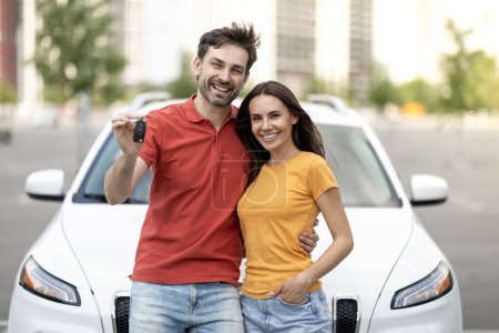Foto de Alegre atractivo millennial hombre y mujer marido y esposa comprar coche nuevo, de pie por el nuevo auto blanco al aire libre en el lugar de estacionamiento, abrazando, mostrando la llave y sonriendo a la cámara - Imagen libre de derechos