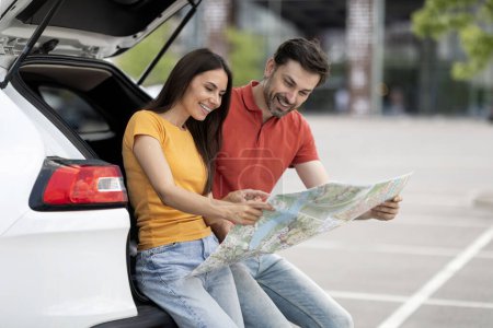 Photo for Cheerful millennial couple friends travelling together by car. Excited young man and woman sitting at auto open trunk, checking map, choosing destinantion to travel. Summer holiday, weekend - Royalty Free Image