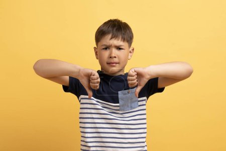 Photo for Unhappy sad cute preteen boy school aged kid wearing tshirt showing thumbs down, expressing disgust isolated on yellow background. Sad child doesnt like something, copy space - Royalty Free Image