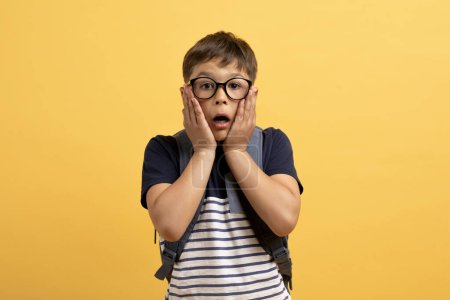 Photo for Amazed cute preteen boy schooler wearing eyeglasses with backpack behind his back touching his face and grimacing isolated on yellow background, kid got stressed before school - Royalty Free Image