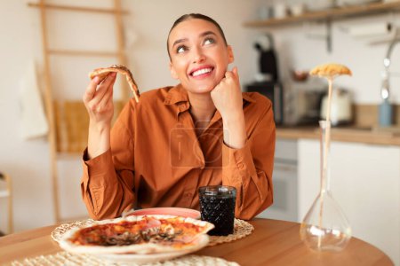 Téléchargez les photos : Jeune femme rêveuse tenant une tranche de pizza savoureuse et rêveuse, assise à table dans l'intérieur de la cuisine, espace libre. Cheat repas, la nutrition et la restauration rapide trop manger - en image libre de droit
