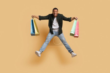 Photo for Full length body size view of attractive cheerful african american guy in casual outfit jumping in the air with new things colorful paper bags in his hands isolated over beige color background - Royalty Free Image