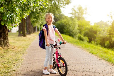 Téléchargez les photos : Fille gaie arrivant à l'école sur son vélo, portant un sac à dos pour le début de l'école, debout près du vélo dans le parc et souriant à la caméra. Concept de retour à l'école - en image libre de droit