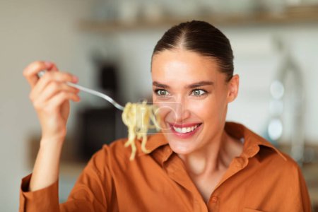 Photo for Hungry young caucasian woman eating tasty homemade pasta, holding and looking at skewered spaghetti on a fork, sitting in kitchen interior background - Royalty Free Image