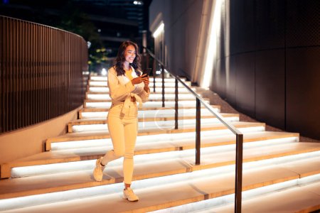 Foto de Mujer joven y elegante sosteniendo el teléfono celular mientras baja las escaleras iluminadas, caminando en la ciudad por la noche, longitud completa, espacio libre. Euforia urbana - Imagen libre de derechos