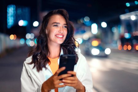 Foto de Hermosa dama caminando por la calle de la ciudad y utilizando el teléfono inteligente en la noche, de pie cerca de la carretera mirando a un lado y sonriendo, espacio libre. El explorador fuera de horario - Imagen libre de derechos