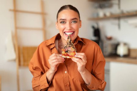 Téléchargez les photos : Femme européenne excitée mordant et appréciant la pizza, tenant une tranche et regardant la caméra, assise à l'intérieur de la cuisine. La malbouffe, la nutrition malsaine triche repas - en image libre de droit