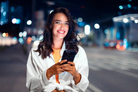 Foto de Paseando por la ciudad nocturna. Retrato de mujer alegre usando el teléfono celular durante la visita nocturna, señora europea mirando y sonriendo a la cámara, espacio libre - Imagen libre de derechos