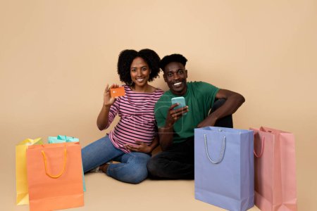 Photo for Cheerful young black couple with belly sit on floor with many packages, enjoy pregnancy use smartphone app and credit card, isolated on beige background, studio. Parents and family, online shopping - Royalty Free Image