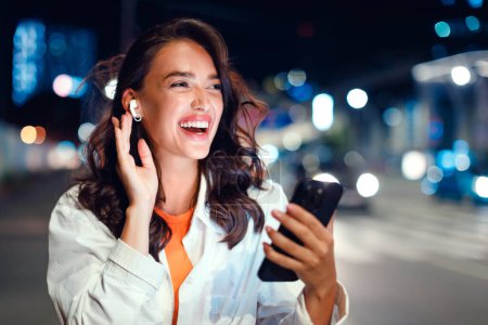 Téléchargez les photos : Des mélodies au clair de lune. Portrait de femme excitée marchant dans la ville la nuit, utilisant un smartphone et écoutant de la musique dans les écouteurs, regardant de côté et souriant, espace de copie - en image libre de droit
